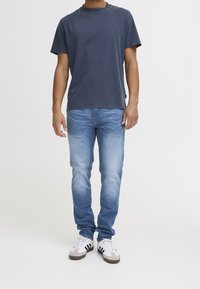 Man wearing a dark blue t-shirt, light blue faded jeans, and white sneakers with black stripes, standing against a plain white background.