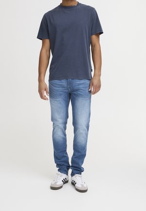 Man draagt een donkerblauw T-shirt, lichtblauwe vervaagde spijkerbroek en witte sneakers met zwarte strepen, staand tegen een effen witte achtergrond.