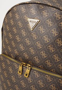 Sac à dos en simili cuir marron avec un logo triangle Guess doré, des motifs embossés, des zips dorés et des détails de surpiqûre contrastants.