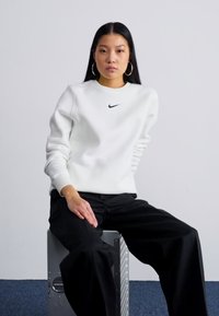 Λευκή φούτερ Nike με στρογγυλή λαιμόκοψη και μαύρο λογότυπο, φορεμένη με μαύρο παντελόνι σε φαρδιά γραμμή, καθισμένη σε μεταλλικό κουτί σε ουδέτερο φόντο.