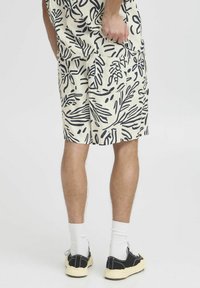 Pantalones cortos beige claros con un patrón de hojas negras, bolsillos frontales y un corte relajado, combinados con zapatos negros y calcetines blancos.