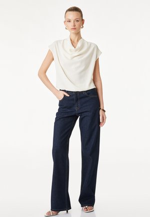 Kvinde iført hvid ærmeløs draperet top og mørke bukser med vide ben, stående med den ene hånd i lommen på hvid baggrund.