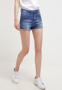 Ljusblå denimshorts med fransad fåll, figurskärning och fem fickor, matchade med vita sneakers och en skjorta med krage.