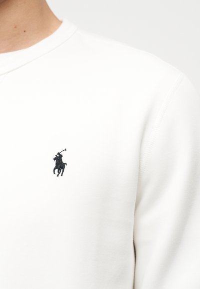 Κοντινό πλάνο ενός ατόμου που φοράει λευκό πουκάμισο με μικρό μαύρο κεντημένο λογότυπο παίκτη πόλο στο στήθος.