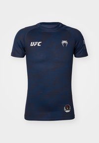 Tamsiai mėlynas sportinis marškinėlis su trumpomis rankovėmis, su tekstūruotomis raštais ir baltu UFC logotipu priekyje ir dešinėje rankovėje. Šešiakampis lopas apačioje.
