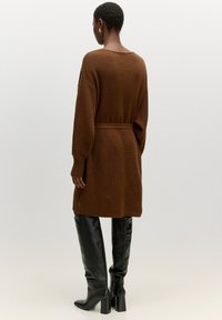 Brauner gerippter Strickcardigan mit langen Ärmeln und einem Bindegürtel, kombiniert mit kniehohen, schwarzen Lederstiefeln mit Absatz. Schlichtes, bequemes Design.