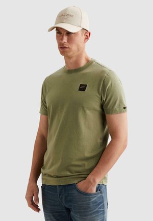 Homme portant un t-shirt kaki à manches courtes avec un petit logo sur la poitrine, un jean bleu et une casquette de baseball beige avec un texte brodé.