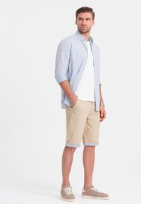 Camicia a righe blu chiaro sopra una maglietta bianca, abbinata a pantaloni corti beige fino al ginocchio e sneakers color taupe. Look semplice e casual.