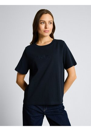 Jeune femme aux longs cheveux bruns portant un t-shirt noir uni à manches courtes et un jean foncé, debout avec les mains derrière le dos.