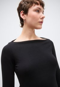 Fekete, hosszú ujjú felső sima anyagból, hajónyakú és testhez simuló szabással, semleges háttér előtt bemutatva.