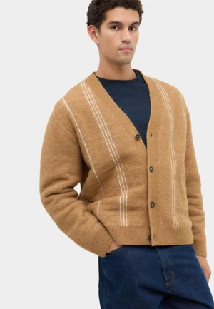 Junger Mann trägt einen beigen, geknöpften Cardigan mit weißen Streifen über einem schwarzen Hemd und blauen Jeans, steht mit einer Hand in der Tasche.