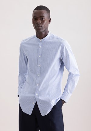 Mężczyzna w jasnoniebieskiej, zapinanej na guziki, pasiastym koszuli z kołnierzykiem typu band i ciemnych spodniach, stojący z rękami w kieszeniach.