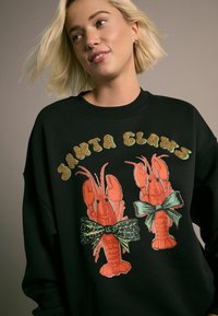 Czarny sweatshirt z żywymi, pomarańczowymi grafikami homarów ozdobionymi kokardkami. Wzór zawiera akcenty brokatowe i ma luźny krój.