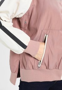 Kevyt pommikonejacka, jossa on vaaleanpunainen vartalo ja valkoiset, vuoratut hihat, joissa on mustat raidat. Sivutaskut ja vetoketjuyksityiskohta helmassa.