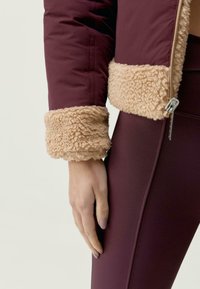 Burgundská bunda s nadýchaným béžovým kožešinovým límcem. Detailní záběr ukazuje hladkou látku a texturované manžety, doplněné pevnými legínami.