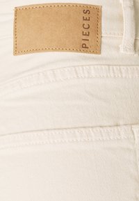 Weißer Denim-Stoff mit einem Lederetikett, auf dem das Wort "PIECES" steht. Enthält gestickte Details und eine glatte, strukturierte Oberfläche.