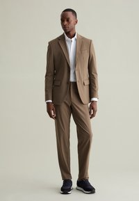 Completo khaki con blazer aderente e pantaloni a gamba dritta, abbinato a una camicia bianca e sneaker blu navy, texture liscia, dettagli minimi.