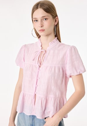 Jeune femme aux longs cheveux porte une blouse rose clair texturée avec des manches bouffantes et de grandes boucles d'oreilles en créoles sur un fond uni.