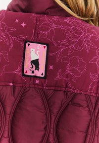 Fialová péřová bunda s kvetinovým vzorom. Obsahuje čiernu nálepku s ružovým dizajnom dvoch mačiek a textom: "Dotknuté mrazom, spálené svetlom."