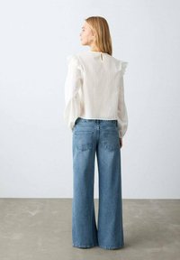 Bílá halenka s volánkovými rukávy a průstřihem u krku, kombinovaná se světle modrými džíny s širokými nohavicemi a zadními kapsami.