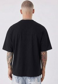 Black cotton t-shirt with short sleeves and a relaxed fit. Notable details include visible tattoos on arms and a silver chain necklace.