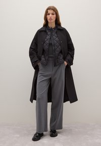 Schwarzer langer Trenchcoat über einer gesteppte schwarzen Jacke, graue hoch taillierte Hose und schwarze Schuhe. Glatter Stoff mit klaren Linien.