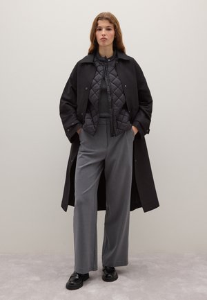 Abrigo largo negro tipo trench sobre una chaqueta negra acolchada, pantalones grises de cintura alta y zapatos negros. Tela lisa con líneas limpias.