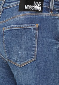Jeans de denim azul con una textura desgastada, que cuentan con un bolsillo trasero y una etiqueta negra con "LOVE MOSCHINO" en letras blancas en la cinturilla.
