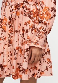 Κοντινό πλάνο χεριού που ακουμπάει σε φόρεμα με λουλουδάτο μοτίβο σε ροδακινί βάση, πορτοκαλί και καφέ λουλούδια, λεπτομέρειες με κουμπιά, ελαστικό μανσέτα και ζώνη στη μέση δεμένη.