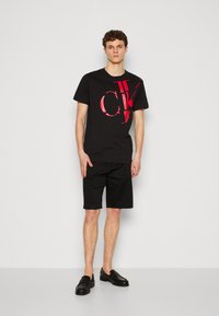 T-shirt en coton noir avec un grand motif rouge "CK", associé à un short noir et des mocassins noirs. Ensemble simple et décontracté.
