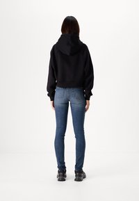 Svart croppad hoodie med huva och sänka axlar, ihop med blå smala jeans. Modellen har på sig svarta sneakers. En enfärgad vit bakgrund.