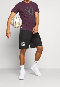 Lila T-shirt med svart NY-logotyp, svarta mesh-shorts med NFL-patch, vita högskurna sneakers, håller en beige basketboll.