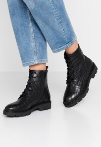 Bottines noires avec une texture crocodile, à lacets sur le devant et fermeture éclair sur le côté. Semelle en caoutchouc robuste et bout arrondi. Portées avec un jean cropped bleu.