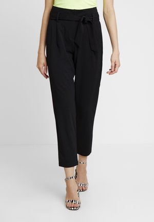 Pantalons noirs taille haute avec un détail de ceinture nouée et des plis, présentant une jambe fuselée. Associés avec des sandales à talons hauts noires et blanches à rayures.