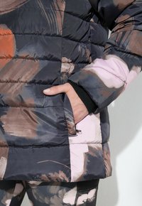 Giacca a vento con un motivo astratto colorato in tonalità scure. Presenta una tasca laterale e polsini elasticizzati, offrendo calore e comfort.