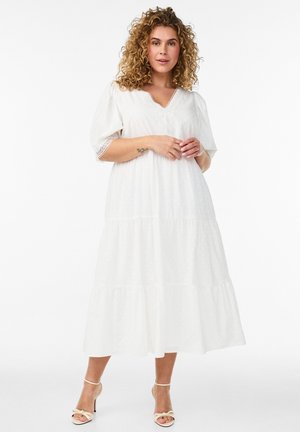 Kvinde med krøllet hår iført en hvid lagdelt midi-kjole med blonder på ærmerne og cremefarvede hæle, stående mod en ensfarvet hvid baggrund.