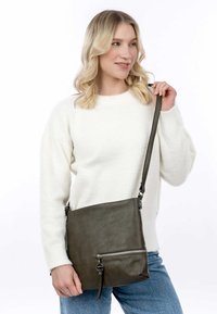 Borsa a tracolla in pelle verde oliva con tasca anteriore con zip, tracolla regolabile e texture liscia, tenuta da una donna in un maglione bianco.