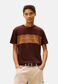 Camiseta de algodón marrón con una franjas horizontal beige que presenta el logo "TOMMYJEANS" en blanco. Diseño de mangas cortas y cuello redondo.