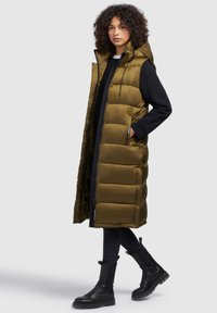 Olive green, long puffer vest with a hood, zip closure, and visible seam stitching. Worn over a black top and paired with dark jeans and black boots.