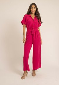 Combinaison fuchsia à manches courtes, devant boutonné, taille ceinturée et jambes larges. Tissu lisse avec une texture subtile. Sandales à talons dorés.