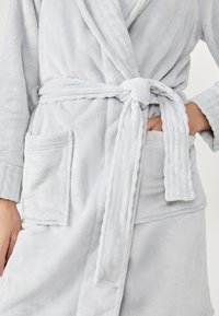 Accappatoio in fleece grigio con texture morbida, colletto a scialle, cintura in vita e tasche laterali. Presenta una finitura soffice e polsini a costine.