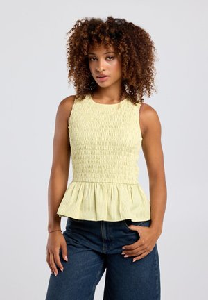 Vrouw met krullend haar, die een gele mouwloze smocked top met peplum en donkerblauwe spijkerbroek draagt, staand met één hand in de zak.