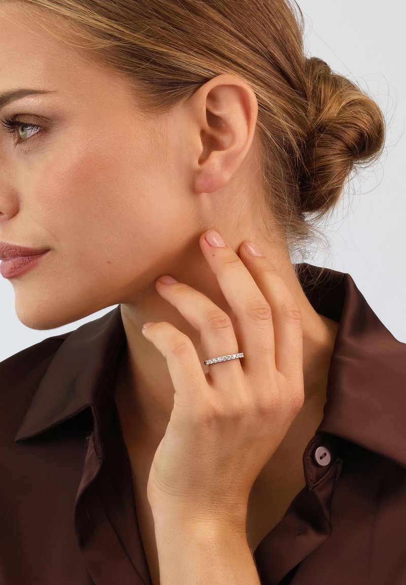Silver ring featuring multiple small clear gemstones, set in a simple band. The model wears a brown satin shirt and touches her neck.