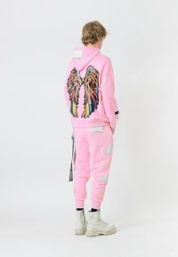 Sudadera rosa con bordado de alas coloridas en la parte de atrás, combinada con pantalones de chándal a juego que cuentan con acentos bordados en blanco y botas blancas gruesas.