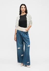 Femme debout, main dans la poche, portant un haut noir, un cardigan blanc rayé, un jean large déchiré et des chaussures blanches pointues sur fond blanc.