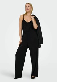 Jumpsuit noir avec des détails en dentelle sur le corps et des fines bretelles, design à jambes larges, associé à un blazer noir. Les accessoires comprennent des boucles d'oreilles et un collier.