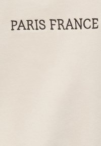Texte brodé noir « PARIS FRANCE » sur fond de tissu beige clair.