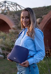 Marineblaue Lederhandtasche mit rundem Design, metallischen Beschlägen und einem Schultergurt, gehalten von einer Person in einem hellblauen Hemd und Jeans.