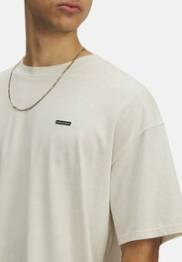 Krēmkrāsas īso piedurkņu T-krekls ar brīvu piegriezumu, uz kura krūšu daļas ir neliels melns Under Armour logo uzplecs, un tas ir ar gludu tekstūru.