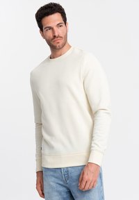 Sweatshirt crème clair avec un col rond, des poignets et un ourlet côtelés. Texture douce. Porté sur un jean en denim bleu. Design simple et minimaliste.